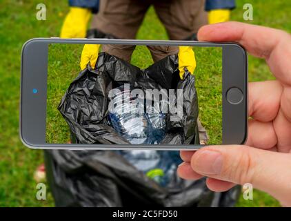 Uomo che tiene un sacchetto di plastica nera con bottiglie di plastica vuote. Sfondo erba. Smartphone fotografico. Smartphone in mano. Plastica sullo schermo. Foto Stock