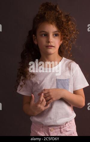 Una bambina in una t-shirt bianca con capelli ricci lunghi, tenendo una palla di decorazione albero di Natale, guarda via su uno sfondo scuro Studio viola Foto Stock