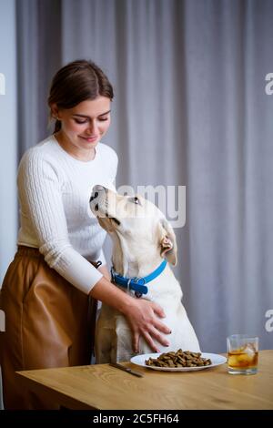 Labrador Retriever giallo è seduto al tavolo con la sua padrona. Cibo cane in un piatto bianco, amico cane mans Foto Stock