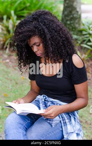 una giovane donna nera tra i 20 e i 30 anni seduta a leggere un libro da sola, in un parco, circondato da alberi Foto Stock