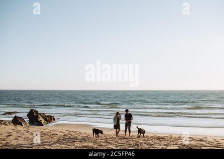 Giovane coppia millenaria con due cani in spiaggia in Portogallo, grandangolo Foto Stock