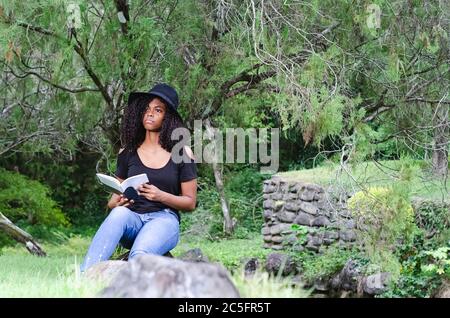 una giovane donna nera tra i 20 e i 30 anni seduta a leggere un libro da sola, in un parco, circondato da alberi Foto Stock