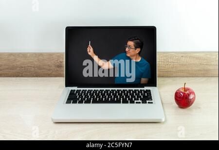 Lezione virtuale insegnata da un insegnante su uno schermo del laptop sorridente mentre scrive su una lavagna nera con una mela sulla scrivania Foto Stock