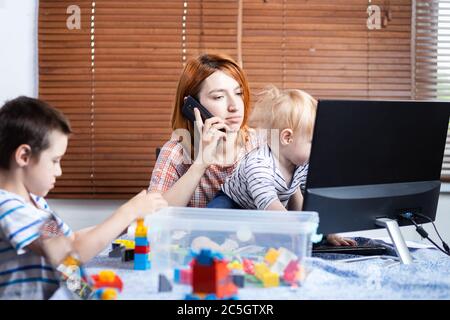 Madre con due ragazzini in ginocchio cerca di ridere a casa . Una giovane donna si prende cura dei bambini e parla al telefono. Lavoro online e bri Foto Stock