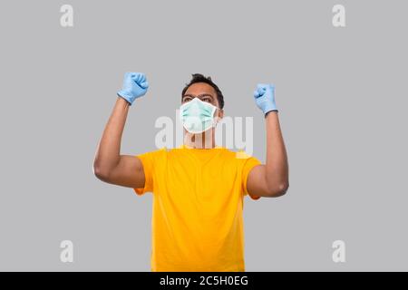 Indian Man molto felice ed eccitato, alzando le braccia, celebrando una vittoria o successo indossando maschera medica e guanti. Firma del vincitore. L'uomo indiano isolato Foto Stock