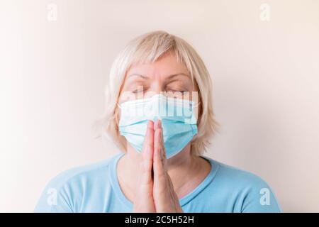 Donna anziana con maschera facciale in preghiera. Ritratto di una donna anziana in una maschera medica che prega. Donna matura che tiene le mani in preghiera. Pregate a casa Foto Stock