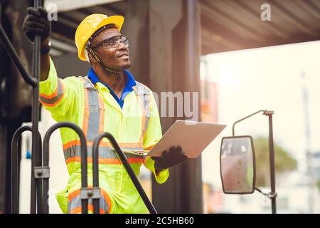 Lavoratore nero afroamericano come caposquadra orgoglioso di felice sorriso di lavoro controllo carico carico carico in magazzino logistica di spedizione con tuta di sicurezza. Foto Stock