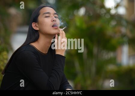 Giovane asiatico con capelli lunghi che fumano al parco Foto Stock