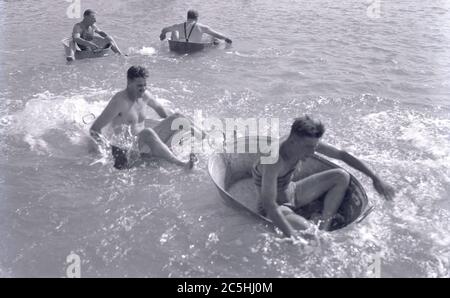 anni '20, storici, uomini in costumi da bagno dell'epoca seduti in vasche di metallo in stagno in una piscina all'aperto, con due di loro che hanno divertente paddstliing in una corsa l'uno contro l'altro. Foto Stock