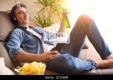 Uomo rilassato che ascolta la musica con le cuffie di un telefono cellulare con una birra in mano seduto comodamente su un divano a casa Foto Stock