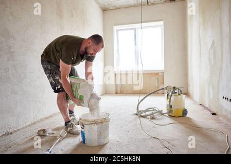 Builder sta andando mescolare stucco in processo di riparazione casa. L'uomo in una piccola stanza preparata per la riparazione versa il composto in un secchio di luce del giorno contro una finestra Foto Stock