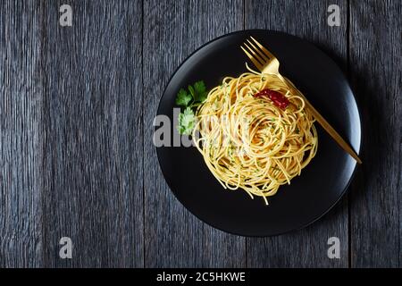 Spaghetti alla colatura di alici, spaghetti con salsa di acciughe, pepe pimento, aglio e prezzemolo su un piatto nero con forchetta dorata su legno scuro Foto Stock
