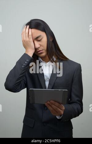 Giovane uomo d'affari asiatico stressato con capelli lunghi che ottengono le notizie difettose mentre usando il tablet digitale Foto Stock