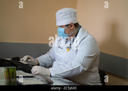 Medico di medicina generale che lavora con il computer in ufficio medico. Medico generico maschile seduto alla scrivania e digitando sulla tastiera. Ospedale cittadino. Maggio 2020 Foto Stock