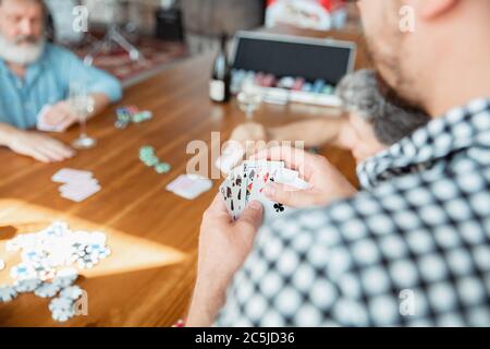 Focus sulle schede. Primo piano gruppo di amici felici e maturi che giocano carte e bevono vino. Guarda felice, eccitato. Uomini caucasici che giocano a casa. Emozioni sincere, benessere, concetto di espressione facciale. Foto Stock