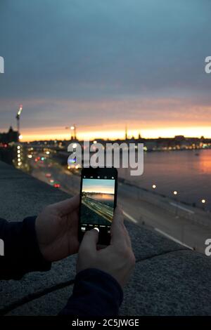 Mani che tengono il telefono cellulare, scattare foto del tramonto in una serata estiva con lo skyline della città vecchia di Stoccolma e l'acqua sullo sfondo Foto Stock