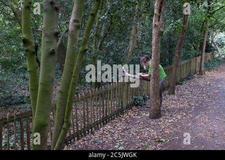 Una signora runner si allunga mentre usa il suo telefono in Holland Park, il 28 giugno 2020, a Londra, Inghilterra. Foto Stock