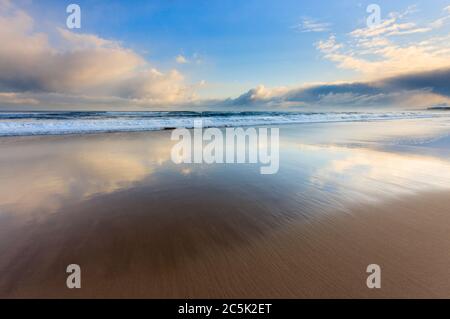 Dopo l'alba tempesta sulla costa, l'ultima delle nuvole minacciose inizia ad ammorbidire la luce e a creare riflessi speculari sulla sabbia della spiaggia. Foto Stock