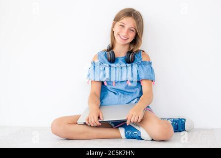 La ragazza sorridente si siede a terra con un computer portatile chiuso e delle cuffie intorno al collo Foto Stock