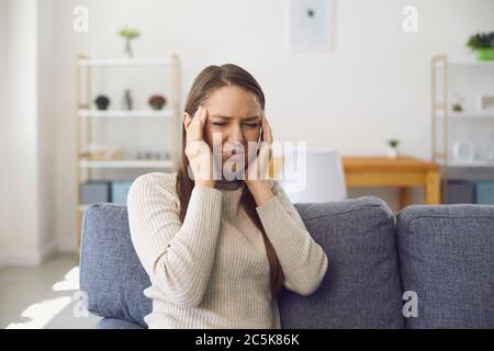 Mal di testa. Emicrania. Una donna bionda ha un mal di testa mentre si siede su un divano in una stanza. Foto Stock