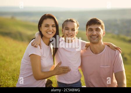 Ritratto di giovane famiglia positiva guardando la fotocamera in campagna. Ragazza carina abbracciando i suoi genitori in vacanza estiva Foto Stock