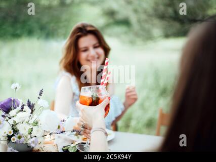 Felice giovane femmina con cocktail alcolici sorridente e che propone toast mentre si siede a tavola durante il banchetto in giardino Foto Stock