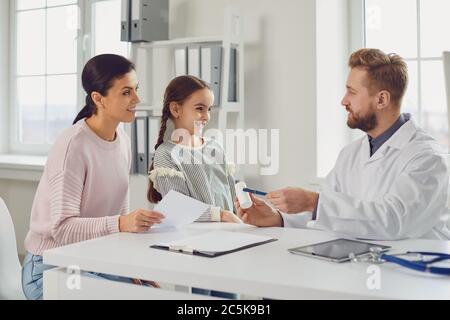 Il medico maschile dà a mamma e figlia una prescrizione mentre si siede a un tavolo in un ufficio clinico. Foto Stock