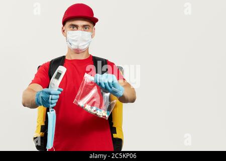 Un corriere in una uniforme rossa, maschera protettiva e guanti consegna pillola farmaco e termometro. Rimani a casa e prenditi cura della tua salute. Corriere del Foto Stock