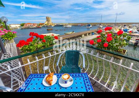 Paesaggio panoramico di gerani rossi fioriti e colazione con croissant e cappuccino in caffetteria italiana. Popolare località balneare Marciana Marina in Foto Stock