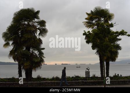 Un pedone è silhoueted mentre cammina tra due palme sul percorso della spiaggia guardando i freighters ancorati in English Bay al crepuscolo Foto Stock