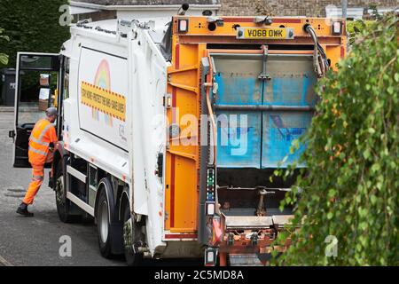 Un raccoglitore di riciclaggio, in un abbigliamento arancione ad alta visibilità, entra in un camion di raccolta rifiuti dopo aver svuotato un bidone di rifiuti nella parte posteriore. Foto Stock