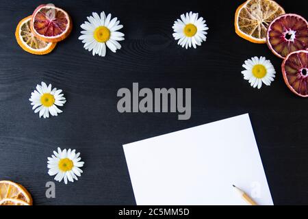 Margherite bianche e arance secche con matita su sfondo di legno scuro. Bella composizione primaverile con carta bianca, modello con spazio di copia per tex Foto Stock