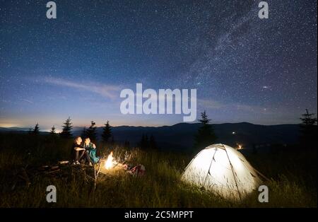 Amici escursionisti seduti su panchina fatto di tronchi, guardando insieme incredibile cielo stellato in montagne accanto a tenda illuminata, cresta di montagna sullo sfondo. Concetto di escursioni, campeggio notturno, relazioni Foto Stock