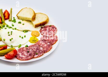 Deliziosa colazione con uova, salumi, cetrioli, pomodori ciliegini, fette di pane, salse e cipolla verde su un piatto bianco. Sano e gustoso m Foto Stock