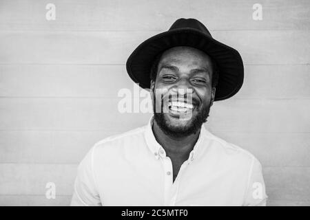 Felice uomo africano ridendo di fronte alla macchina fotografica mentre indossa cappello alla moda - Ritratto di giovane persona nera che si diverte all'aperto - Focus on Face - Nero e. Foto Stock