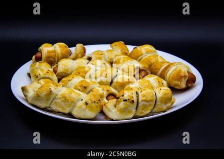 Gli ausages cotti nell'impasto su un piatto bianco su sfondo nero. Salsicce Puff Pasticceria Bunds. Avvolte in pasta sfoglia e cotta sul piatto. Foto Stock