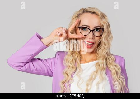 Ritratto di una donna bionda affascinante e simpatica in occhiali che si inocchiano felicemente, mostrando un gesto di vittoria della pace come un'emozione ottimista e ingannatrice Foto Stock