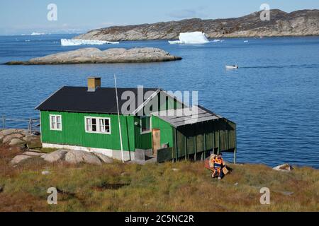 Il lavoratore vestito con colori brillanti raccoglie la spazzatura da una piccola casa verde arroccata sulle rocce che è dwarfed da enormi iceberg che galleggiano. Foto Stock