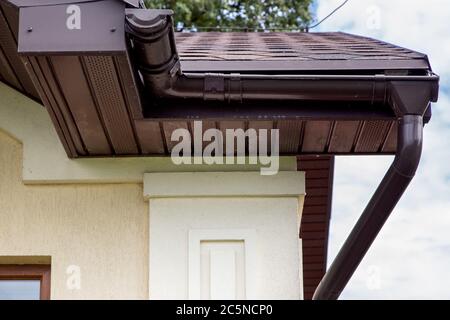 grondaia di drenaggio in plastica marrone su un tetto con piastrelle bituminose vicino ai tubi di drenaggio della casa. Foto Stock