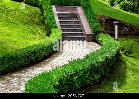 scala in pietra con un passaggio pedonale in piastrelle circondato da cespugli con una siepe con un prato verde in un parco collinare, nessuno. Foto Stock