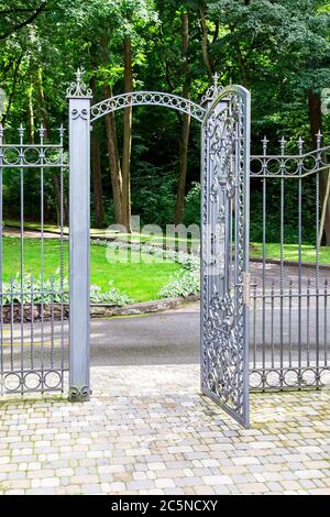 cancello di ferro nella recinzione ingresso al cortile, una barriera tra il parco con piante e il marciapiede fatto di piastrelle. Foto Stock