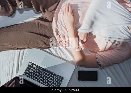 Donna con braccio ferito si appoggia a letto accanto a un computer e telefono. Concetto di lavoro remoto, non essere in grado di essere produttivo, rimanere in un congedo malato Foto Stock