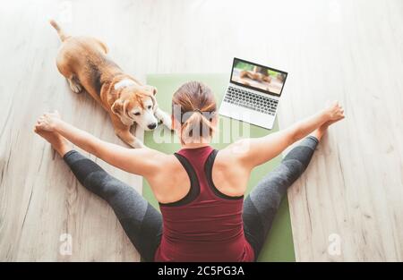 Vista dall'alto in forma sportiva donna sana seduta sul tappetino in Upavistha Konasana posa, facendo esercizi di respirazione, guardando online yoga classe sul computer portatile. Foto Stock