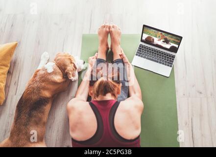 Vista dall'alto in forma sportiva donna sana seduta sul tappetino in posa Paschimottanasana, facendo esercizi di respirazione, guardando una lezione di yoga online sul computer portatile. H Foto Stock