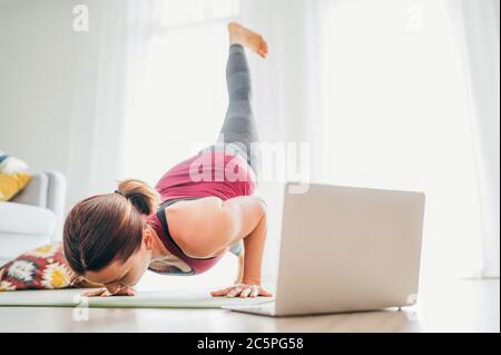 Adatta donna sana sportiva sul tappetino in Eka Pada Chaturanga Dandasana personale a quattro zappi posa, facendo esercizi di respirazione, guardando online yoga classe sul lapt Foto Stock