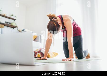 Adatta donna sana e sportiva sul tappetino in posa Cat Majaryasana, facendo esercizi di respirazione, guardando una lezione di yoga online sul computer portatile. Persone sane e. Foto Stock