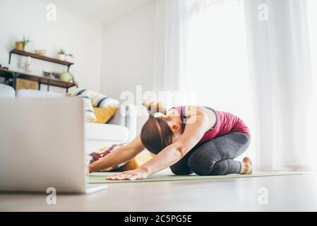 Calzare donna sana e sportiva sul tappetino in Balasana di bambino posa yoga rilassante, facendo esercizi di respirazione, guardando una lezione di yoga online sul computer portatile Foto Stock