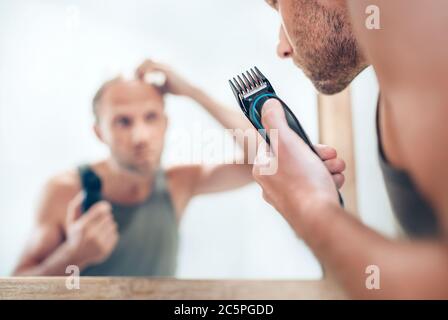 Uomo che guarda lo specchio del bagno e pensa al suo nuovo stile di taglio dei capelli che analizza i capelli.rifinitore elettrico ricaricabile primo piano immagine messa a fuoco. De radicale Foto Stock