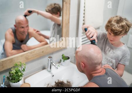 Figlio adolescente aiutando suo padre a tagliare delicatamente una testa calva usando il rifinitore elettrico ricaricabile in bagno. Divertente casa scena e parenti conc Foto Stock