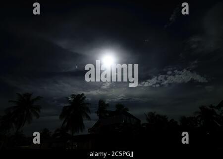 Una notte di luna piena in un villaggio con nuvole sopra e gli edifici e gli alberi sotto con alone che circonda la luna Foto Stock